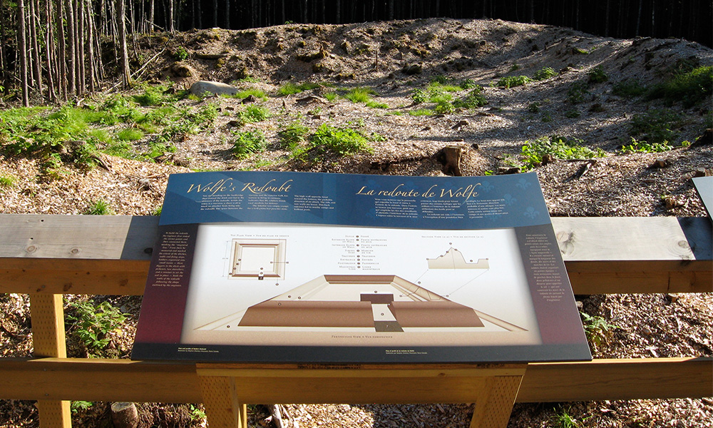 Photograph of panel five, with the redoubt illustration, installed at the Fortress of Louisbourg National Historic Site, directly in front of the redoubt remains.