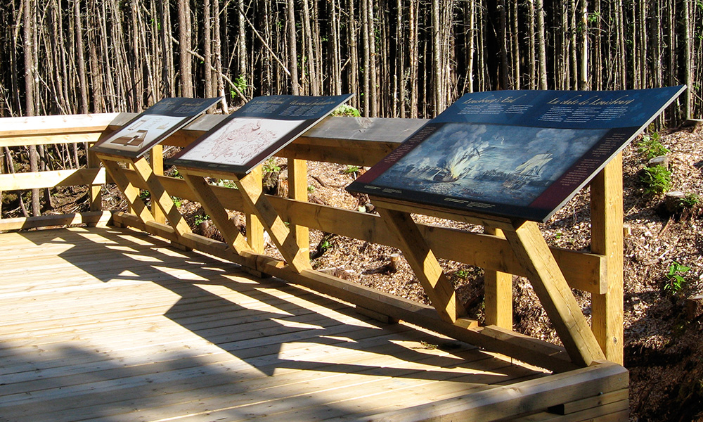 Photograph of panel five, with the redoubt illustration, installed at the Fortress of Louisbourg National Historic Site, directly in front of the redoubt remains.